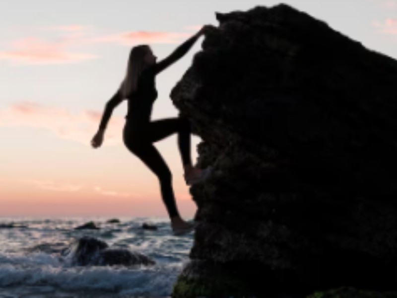 mujer escala una montaña con sus manos, al fondo el atardecer